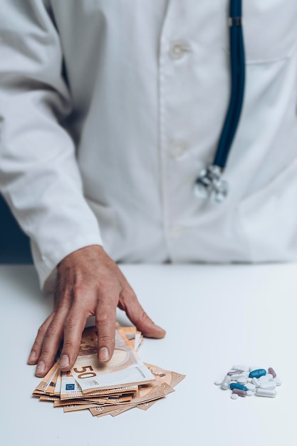 Hand of doctor taking bunch of bills next to bunch of pills on white table