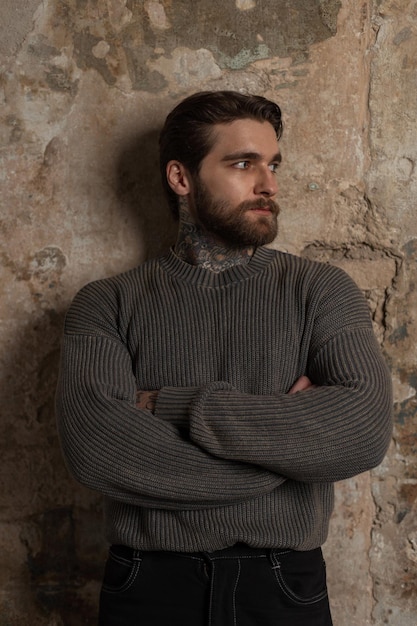 handsome brutal man model with a beard and hairstyle in a fashion vintage knitted sweater stands near a grunge concrete wall