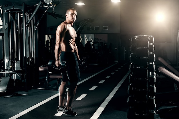 Handsome guy of European appearance, bodybuilder, stands in the gym with dumbbells in his hands. The concept of sports training, fitness training, workout in the gym.