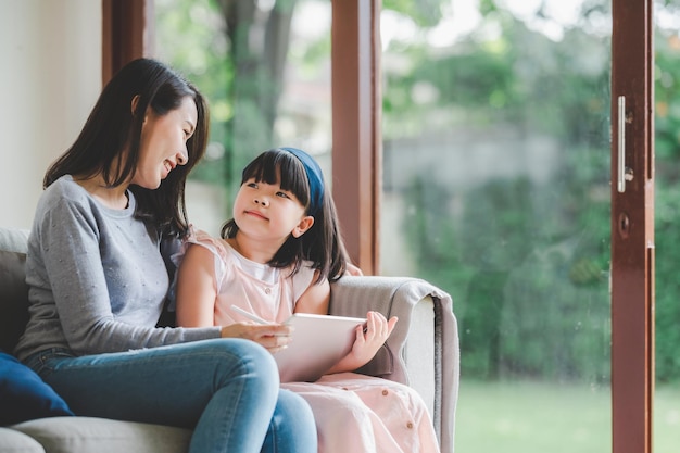 Happy Asian family mother and daughter using digital tablet to study together at home photo focused on mother