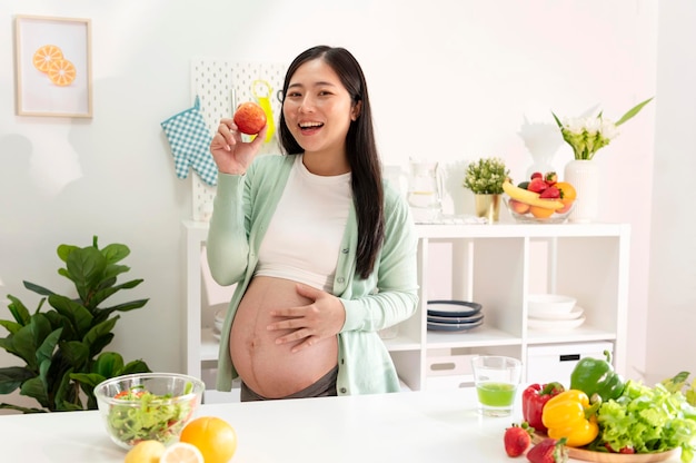 Happy asian pregnant woman eating apple in kitchen at home eating many different fruit during pregnancy healthy pregnancy concept