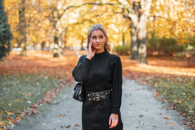 Happy beautiful fashionable blond girl with a black fashion vintage knitted dress with a handbag walks in the autumn park