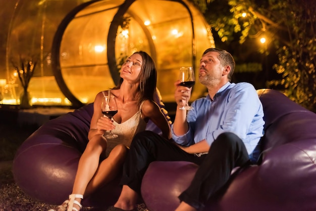 A happy couple drinking wine near a fireplace at night Transparent bubble tent on the background