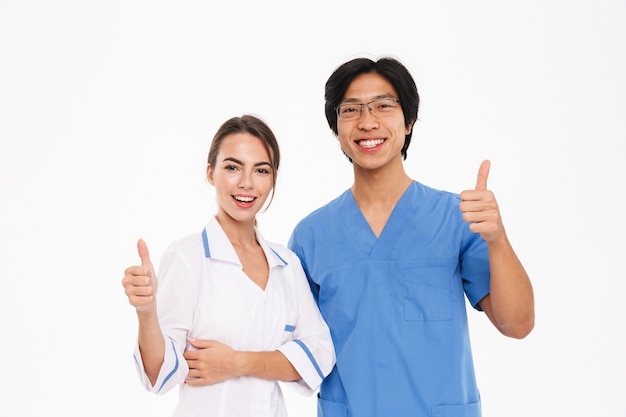 Happy doctors couple wearing uniform standing isolated over white wall, showing thumbs up