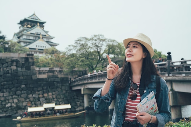 Photo 幸せな女性のアジア人観光客が、ガイドブックと紙の地図を路上で指さしている。石の橋の下の背景にある川湖の木竹イノシシ。強力な壁日本に囲まれた大阪城。