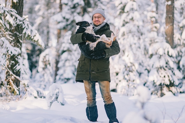 Happy man holding lovely dog in his hands in snowy forest. Smiling boy hugging adorable puppy in winter wood. Pet lover.  Dog - human`s friend concept.