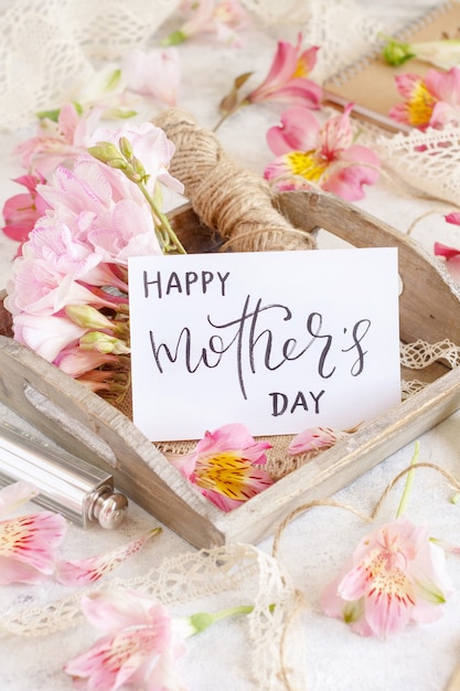 Happy Mother's Day handwritten card on a wooden tray between pink flowers close up