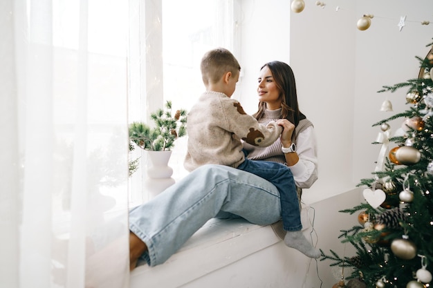 Happy mother with son sit on windowsill near christmas tree