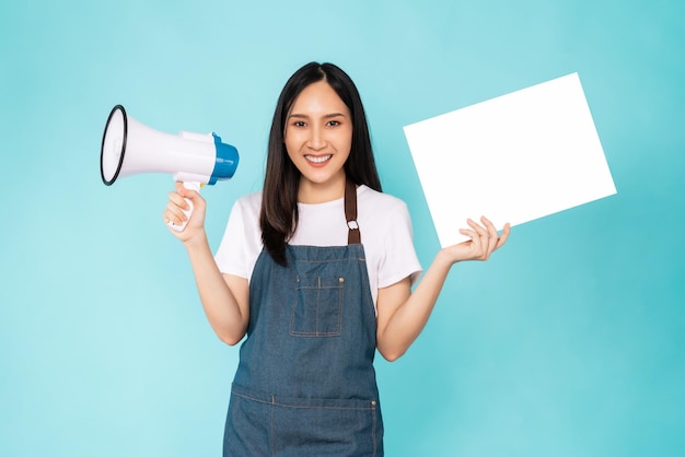 Happy young Asian woman wears an apron and holding megaphone with showing a blank paper on blue background