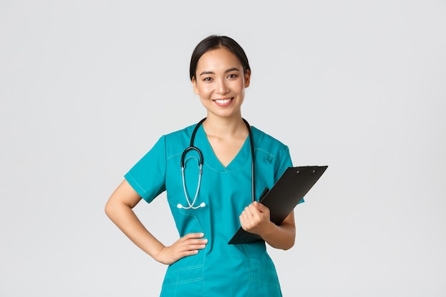 Healthcare workers, preventing virus, quarantine campaign concept. Smiling pleasant asian female physician, doctor during examination wearing scrubs and holding clipboard, white background.