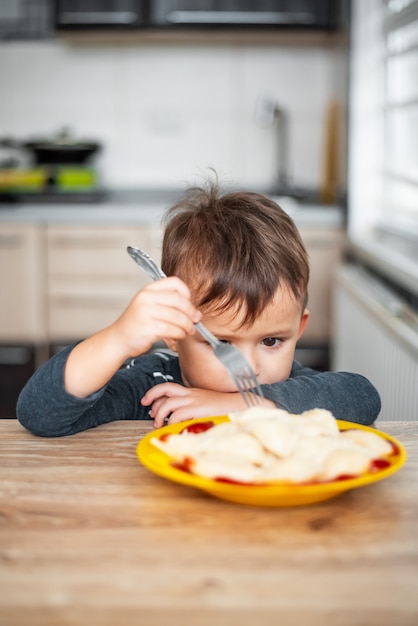 Hungry child eating dumplings in the kitchen, sitting at the table in a gray jacket