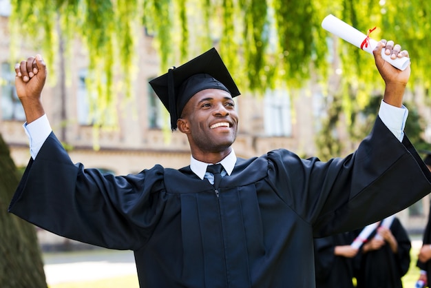 I have finally graduated! Happy young African man in graduation gowns holding diploma and rising arms up while his friends standing in the background