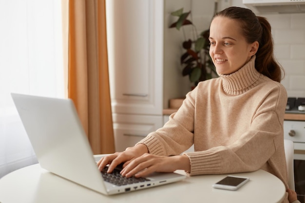 Image of woman with dark hair wearing beige sweater sitting in kitchen and working on laptop and typing on keyboard expressing positive emotions