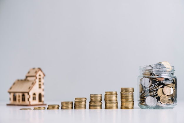 Increasing stacked coins and glass jar in front of house model