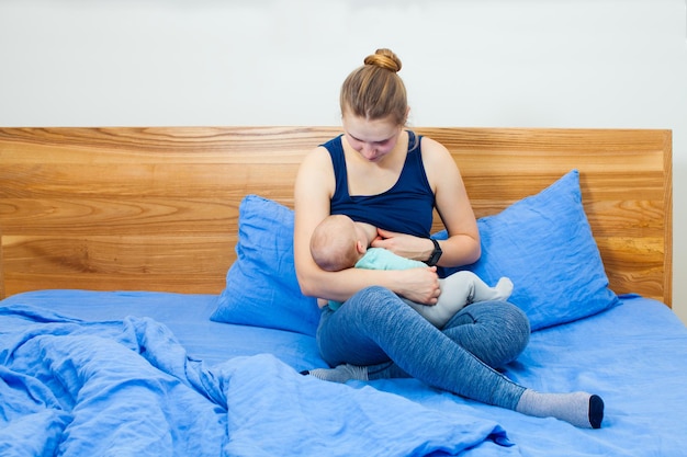 Jonge moeder zittend in haar bed, comfortabel ondersteund door kussens, haar baby in wiegpositie op schoot, borstvoeding. Goede positie voor succesvolle borstvoeding