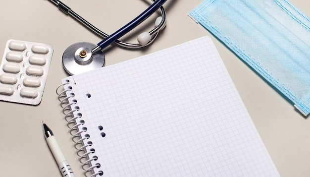 On a light gray background, a medical mask, a stethoscope, pills and a notebook with a pen. Medical concept