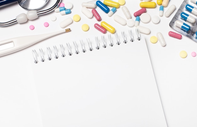 On a light surface, multi-colored pills, a stethoscope, an electronic thermometer and a blank notebook with a place to insert text