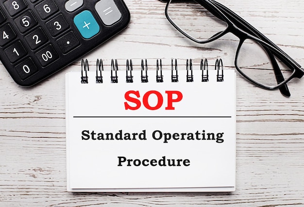 On a light wooden table calculator glasses and a blank notepad with the text SOP Standard Operating Procedure Business concept