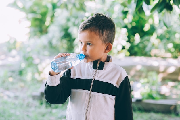 A little boy drinks water outdoors from a blue bottle