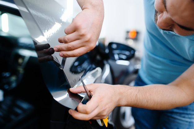 Male wrapper installs car tinting, tuning service. Worker applying vinyl tint on vehicle window in garage, tinted automobile glass