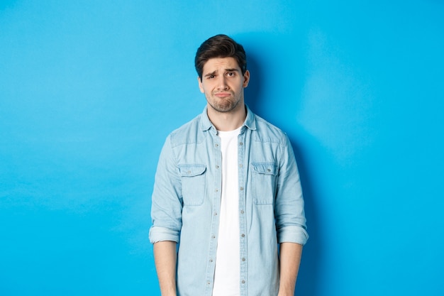 Man looking with doubt at front, frowning and sulking, feeling uneasy about something, standing against blue wall