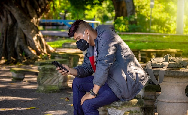 Man with a mask sitting on a bench with his phone young man with a mask sitting with a cell phone