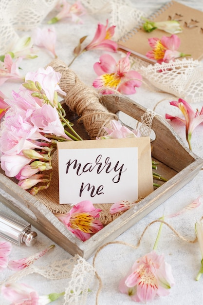 MARRY ME handwritten card on a white table between pink flowers close up