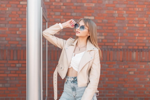 Mooie zoomer modieuze jonge vrouw model in een witte leren jas met een top en jeans draagt een zonnebril in de buurt van een bakstenen muur in de stad Stedelijke vrouwelijke jeugd streetstyle kleding look