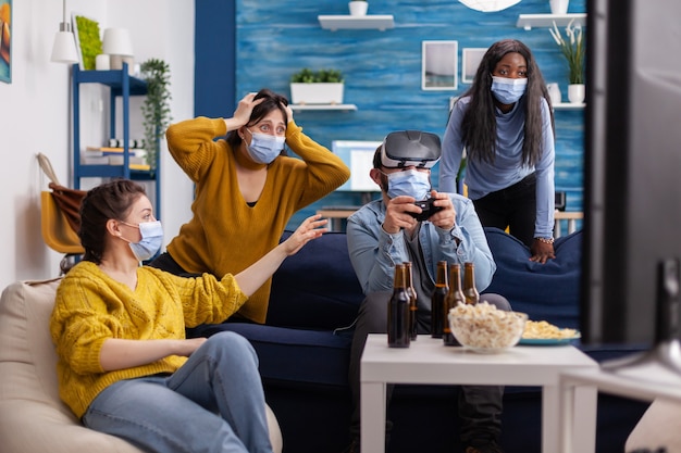 Multiethnic friends enjoying vr tehnology playing video games in living room wearing mask to prevent infection with coronavirus keeping social distancing. Diverse people having fun at new normal party