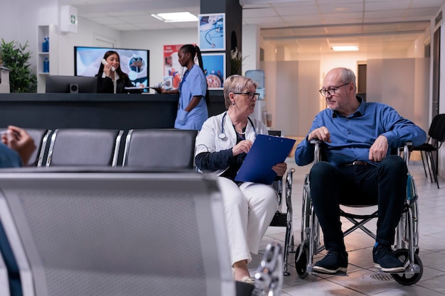 Old doctor consulting man with disability in waiting room lobby, doing examination appointment with wheelchair user. Specialist and patient with physical impairment talking about healthcare support.