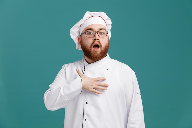 Onder de indruk jonge mannelijke chef-kok met een uniforme bril en een pet die naar de camera kijkt en de hand op de borst houdt geïsoleerd op blauwe achtergrond