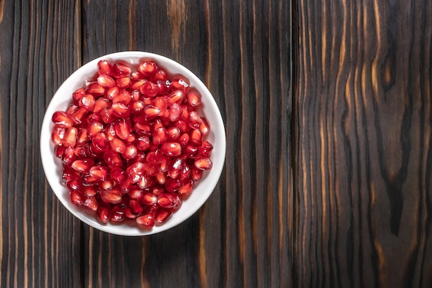 Pomegranate seeds heap in white plate on dark wooden background with place for text, top view