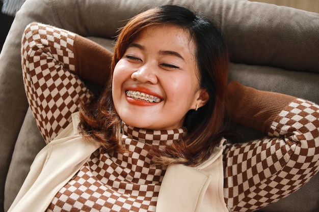 Photo portait of enjoying asian adult woman wear braces showing relax on sofa with hands