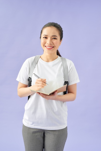 Photo portrait of a happy casual girl student with backpack writing in a notepad while standing with books isolated over purple background