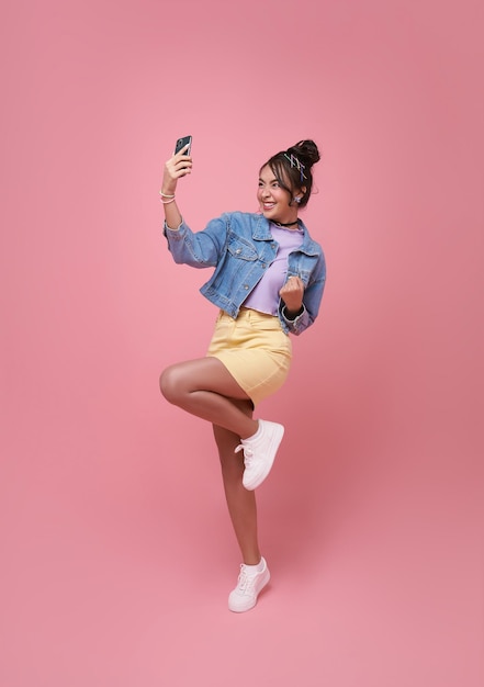 Portrait of a happy young asian woman celebrating with mobile phone isolated over pink background