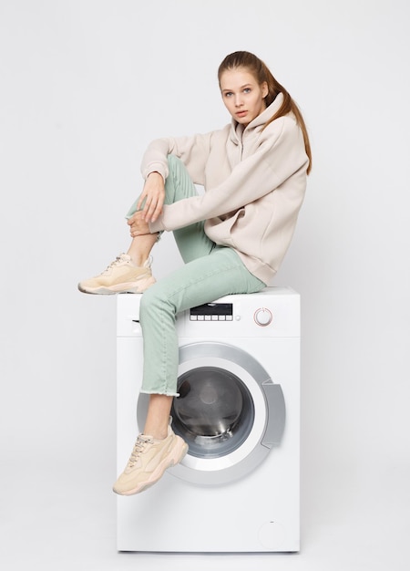 Portrait of young beautiful young girl sitting on a washing machine and looking at the camera isolated on white background