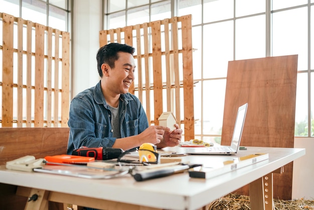 Foto portret van glimlach aziatische mannelijke vader die een klein houten huis bouwt en bij elkaar houdt in de houtbewerkingsstudio thuis met het meetinstrument en werkgereedschap op tafel