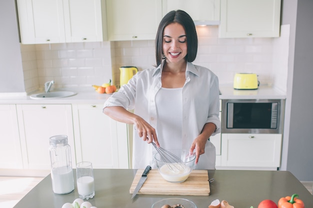 Positive and nice girl stands in kitchen at table and blending together milk with eggs.