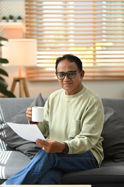 Positive senior man holding paper make loan insurance payment on laptop while sitting in cozy living room Elderly technology concept