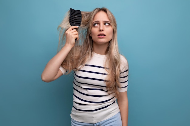 Sad upset blond young woman with difficulty combing her hair that falls out on a blue background