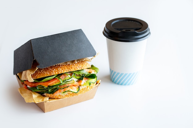 Sandwich with red fish, cheese, arugula, avocado and cucumber.Burger with fish with you.Takeaway food.Home delivery food.The sandwich is packed with coffee.Coffee and takeaway.Stylish food packaging