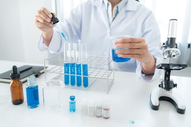 Scientist woman holding beaker and dropper to dropping blue liquid into test tube while working with microscope to analyzing and developing coronavirus vaccine in laboratory