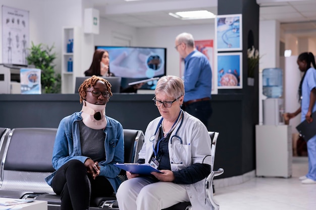 Senior medic consulting woman at facility, wearing cervical neck collar to cure injury and pain. Specialist and patient with medical foam brace talking about recovery in waiting room at health center.