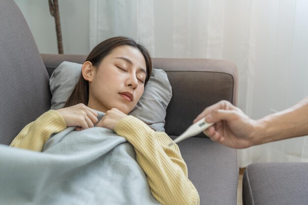 Photo sick, influenza asian young woman, girl headache have a fever, flu and check thermometer measure body temperature, feel illness sitting on sofa bed at home. health care person on virus, covid-19.