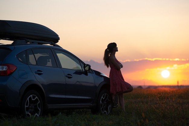 Silhouette of female driver standing near her car on grassy field enjoying view of bright sunset Young woman relaxing during road trip beside SUV vehicle