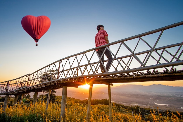 Silhouetted of Asian man standing on Bamboo bridge with Red hot air balloon in the shape of a heart over the  sunset.