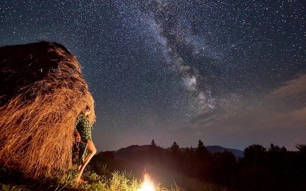 Silhouettes of girls near haystack and bonfire under starry sky on which Milky Way is visible