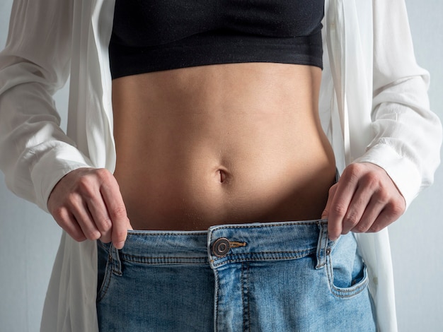 A slender woman with a bare stomach shows how she lost weight. Jeans have become too big. The concept of diet and healthy eating