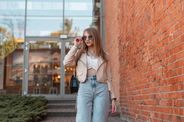 Stylish beautiful trendy woman in a fashionable leather jacket with vintage high waist jeans and a bag wears sunglasses and walks near the shopping mall in the city