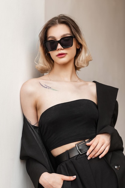 Stylish beautiful vogue elegant business model girl with fashionable sunglasses in fashion black business clothes with top and blazer standing near the white wall Women's street portrait of woman